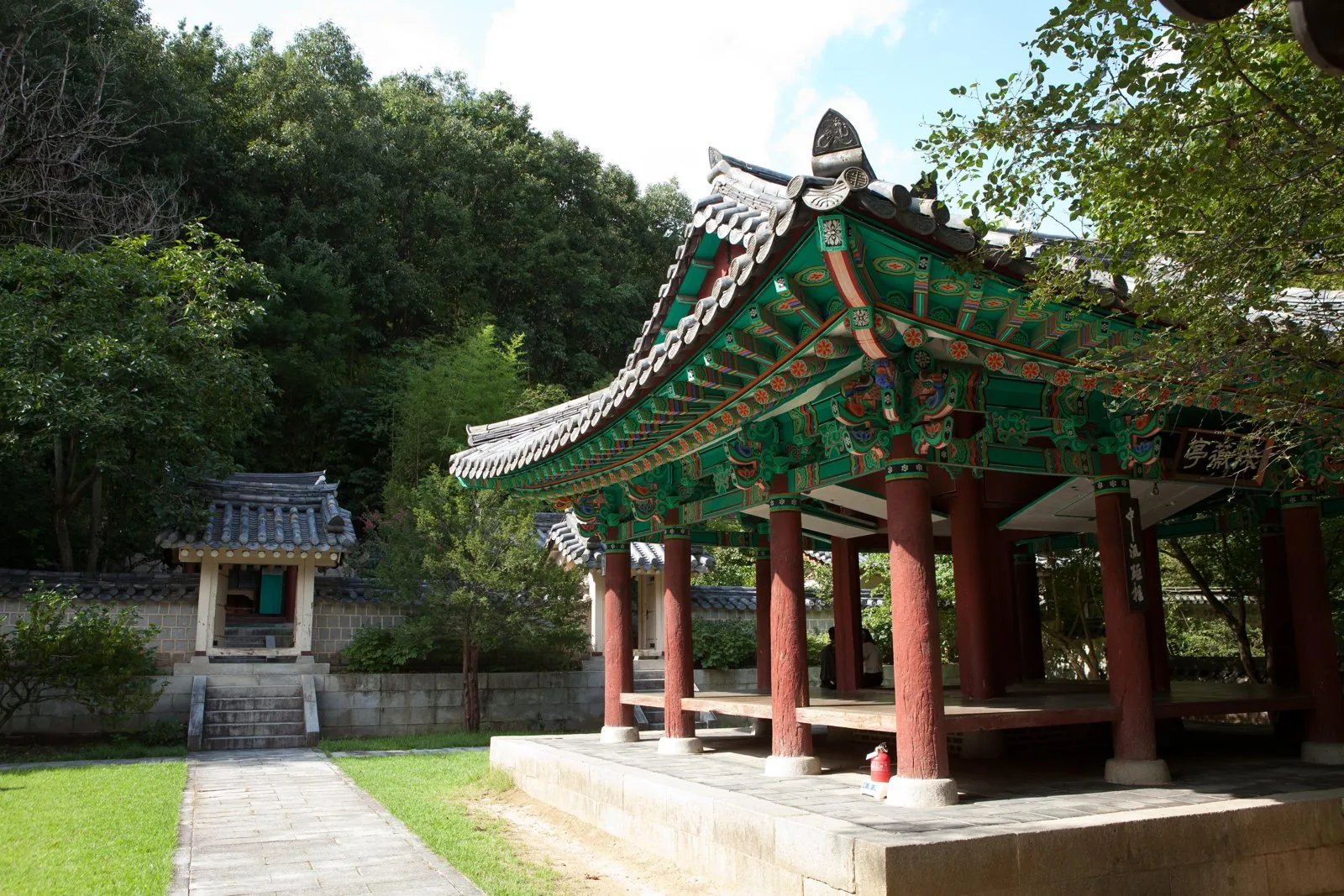 여름, 초록 숲 앞에 선 채미정의 옆모습. 팔작지붕 끝과 단청 처마, 적색 기둥이 보이고, 왼쪽에 작은 문과 돌계단이 있다.